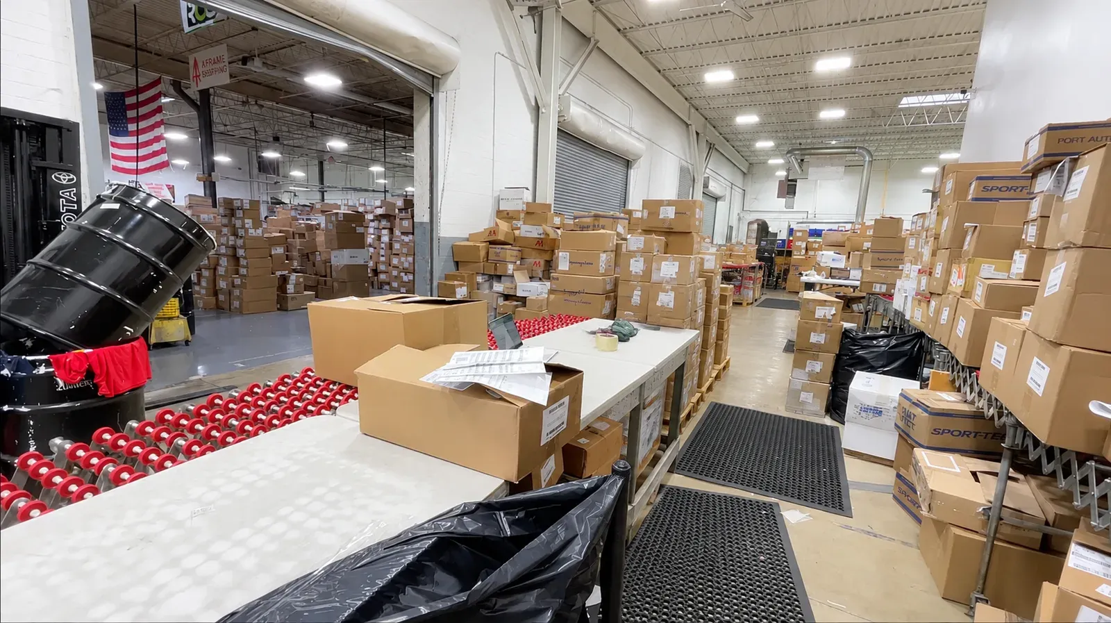 Apparel Redefined receiving dock, stacked wall to wall with cartons of blank garments on pallets and shelves, a roller conveyor and a packing table in the foreground.