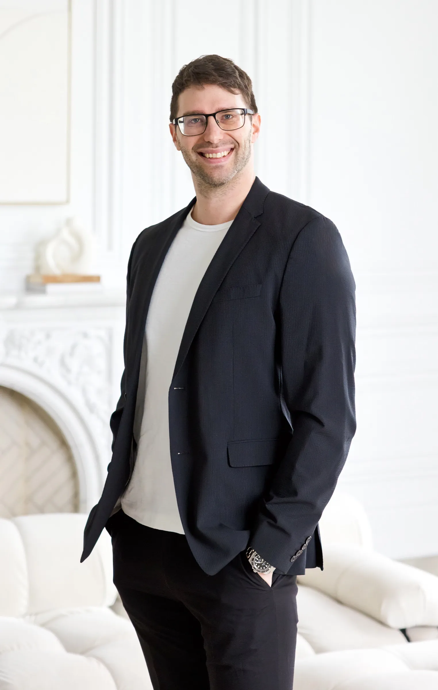 Derek Wayne standing in a bright white room, smiling, in a dark blazer over a white t-shirt.